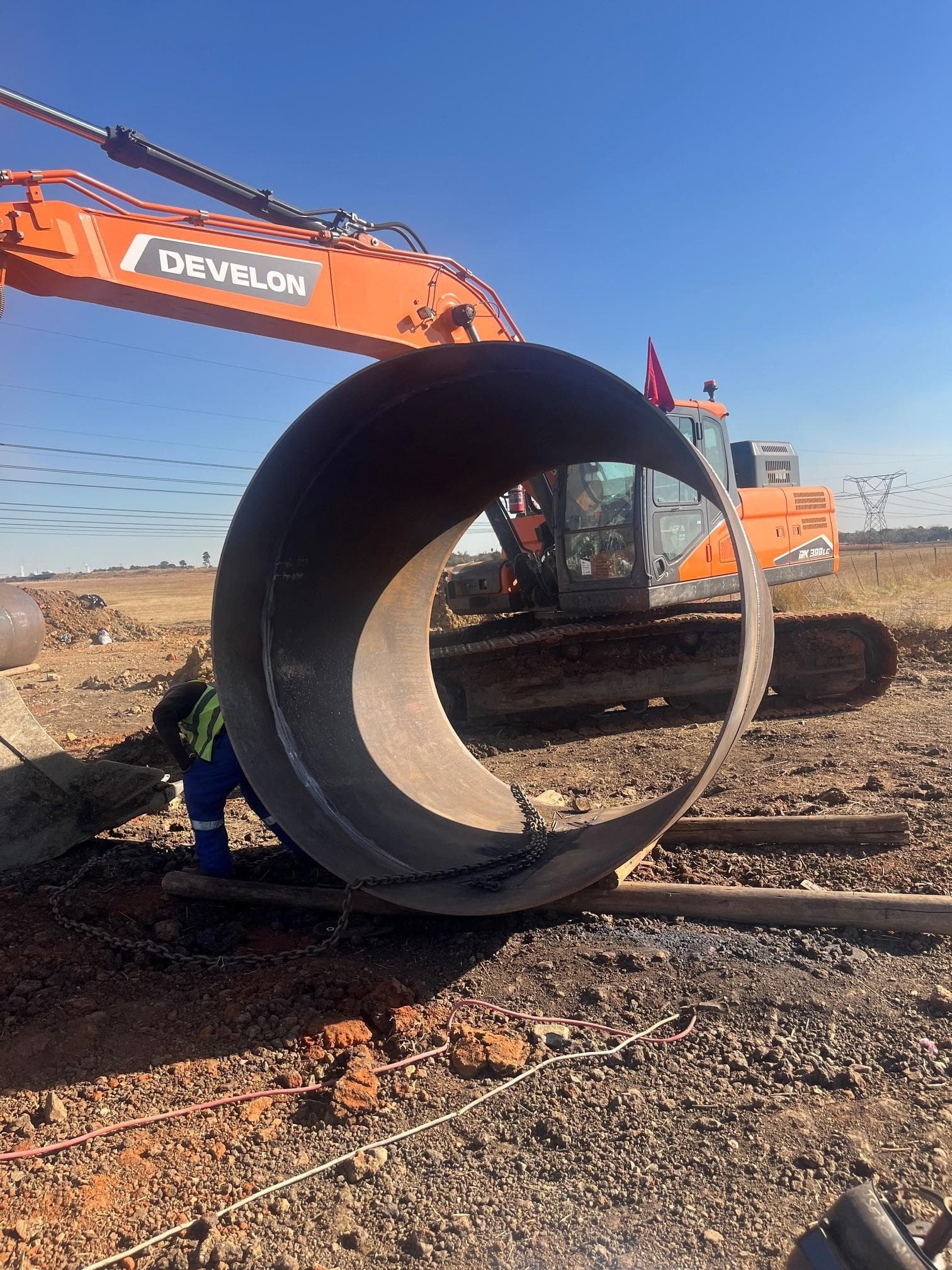 Steel pipe section being prepared for lift beside a Develon excavator on Crosby